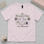 A pale mauve short-sleeve t-shirt lays flat on a grey cutting mat with quilting accessories in the corners. The graphic on the t-shirt features the words 'Measure Once, Cut Twice, Swear Occasionally' around patchwork stars and quilting tools.
