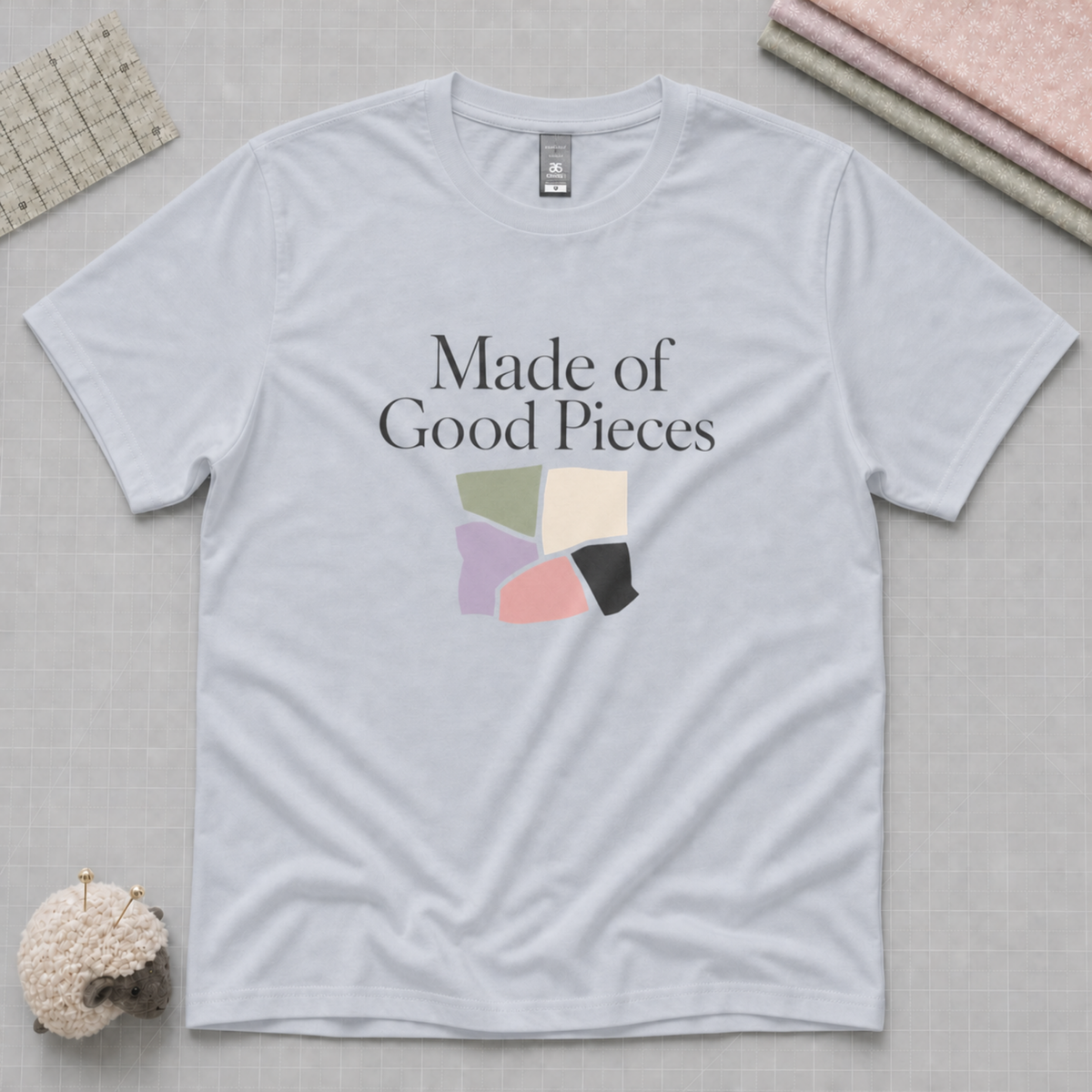 A pale powder blue crewneck t-shirt with the text 'Made of Good Pieces' and a colorful geometric patchwork graphic on the chest, displayed in a flat-lay arrangement with sewing and crafting props.