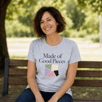 A woman with short brown hair sits smiling on a wooden park bench, wearing a light powder blue t-shirt with the text 'Made of Good Pieces' and a colorful geometric graphic.