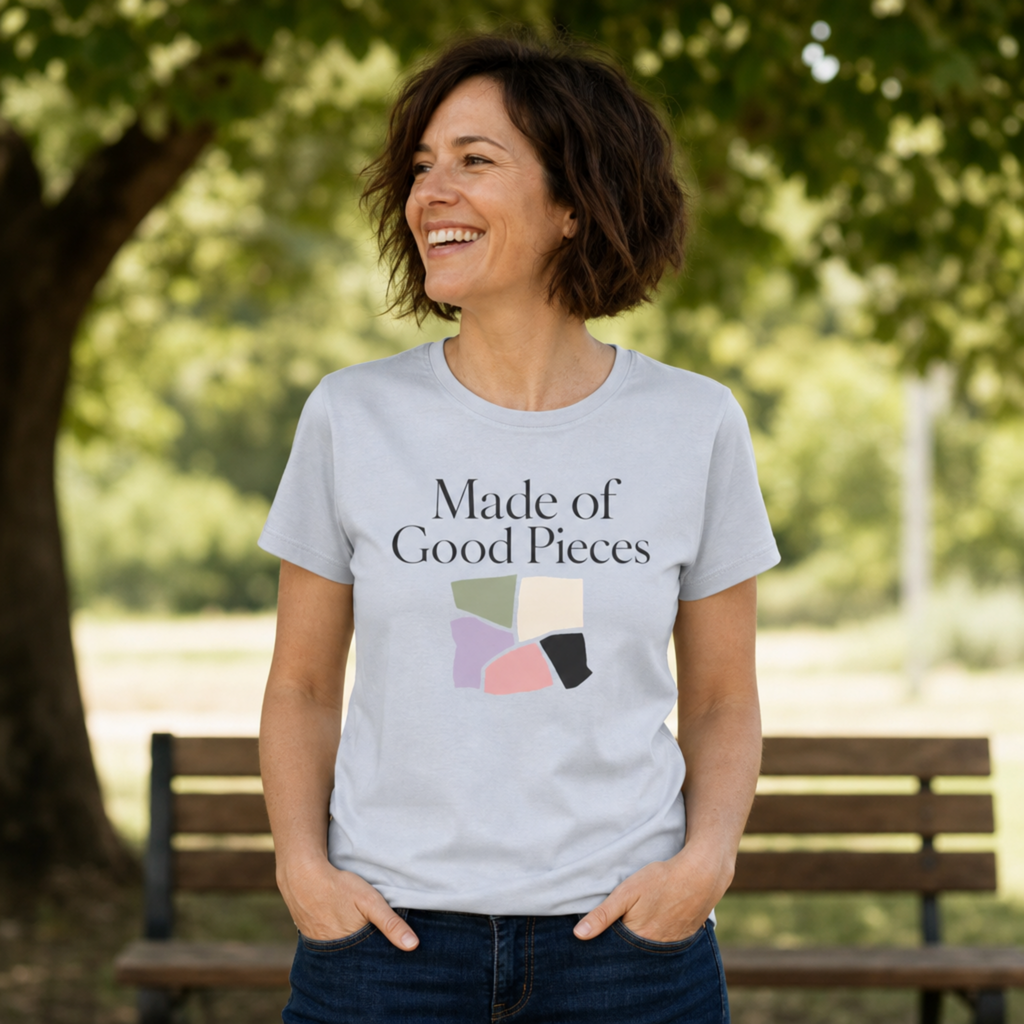 A woman wearing a light powder blue graphic t-shirt with the text 'Made of Good Pieces' and a colorful geometric design, standing outdoors in front of a park bench and trees.