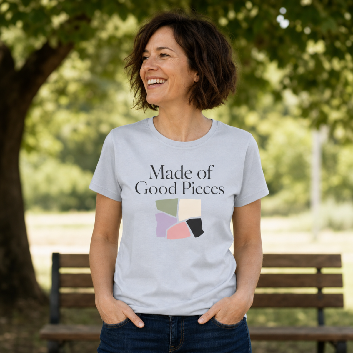 A woman wearing a light powder blue graphic t-shirt with the text 'Made of Good Pieces' and a colorful geometric design, standing outdoors in front of a park bench and trees.