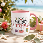 A white ceramic mug with a pink handle and interior, printed with the phrase ‘Merry Stitchmas' in black and red lettering decorated with holly, a purple bobbin, pink pincushion and green thread spool. The mug is styled on a wooden surface with eucalyptus leaves, a starfish, a pink flower, and a blurred beach and Christmas tree background.