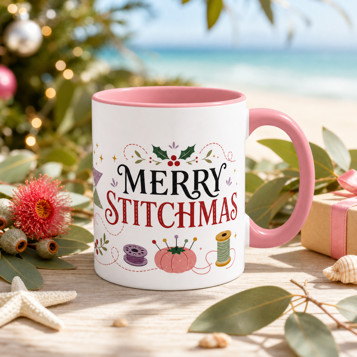 A white ceramic mug with a pink handle and interior, printed with the phrase ‘Merry Stitchmas' in black and red lettering decorated with holly, a purple bobbin, pink pincushion and green thread spool. The mug is styled on a wooden surface with eucalyptus leaves, a starfish, a pink flower, and a blurred beach and Christmas tree background.