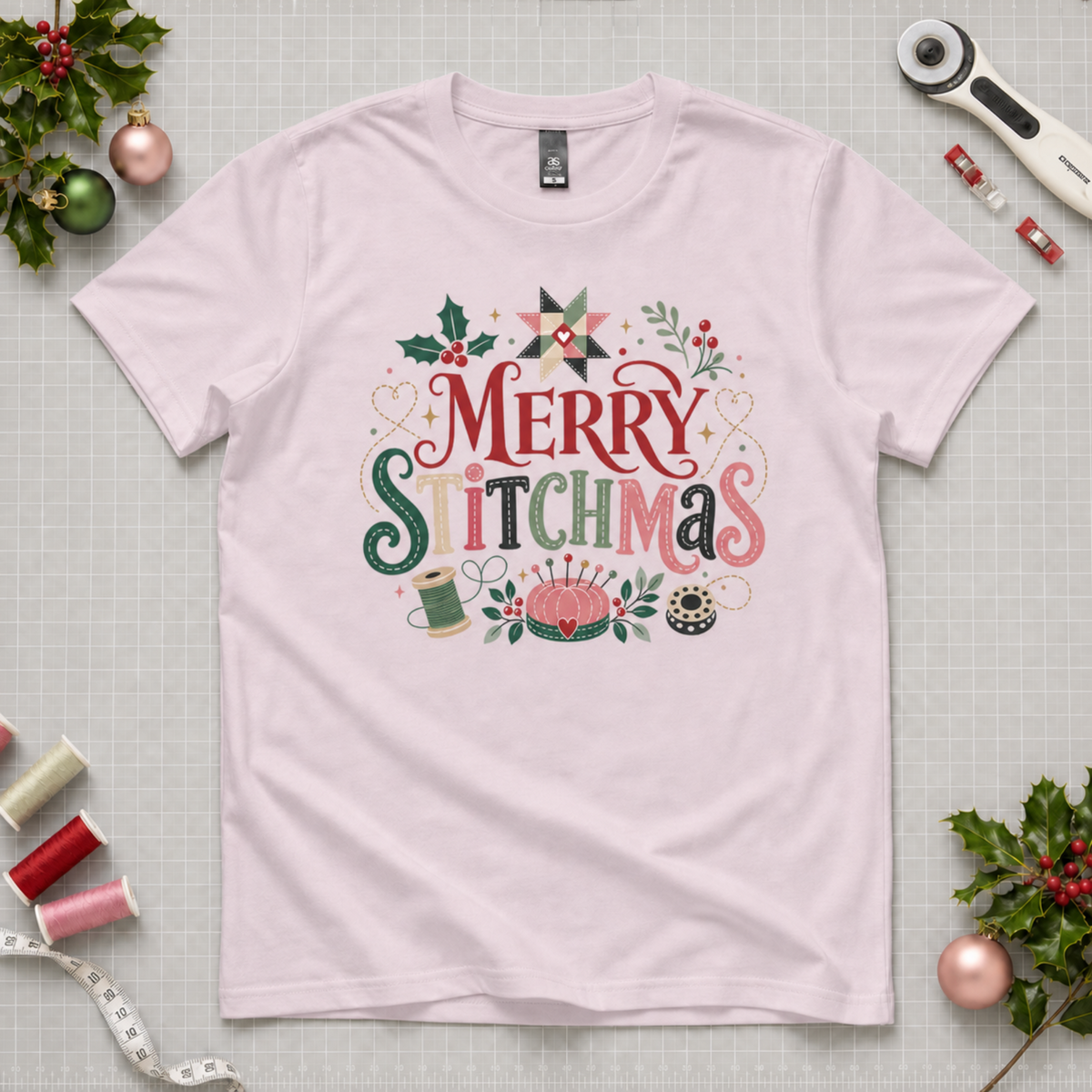 A graphic pale mauve crewnect, short-sleeve tshirt laying flat on a pale grey cutting mat with Christmas decorations and sewing equipment in the corners. The graphic reads 'Merry Stitchmas' surrounded by holly, s quilt star, thread spool, pincushion and bobbin.