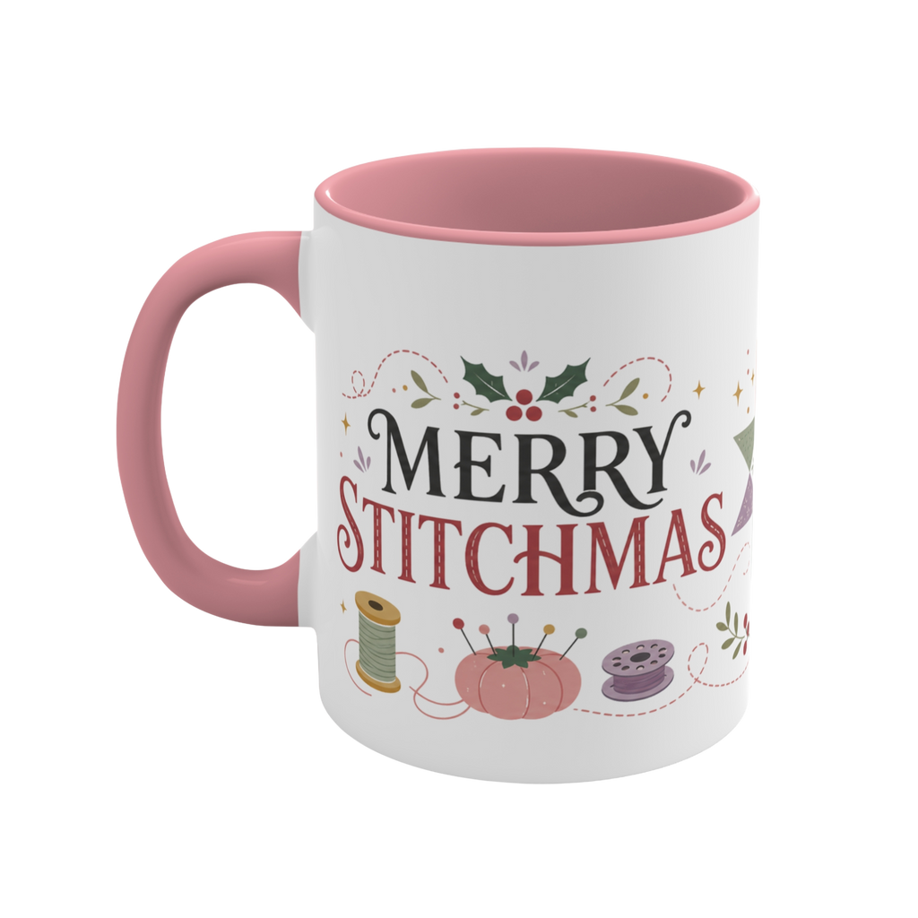 The left side of a white ceramic mug with a pink handle and interior, decorated with a bobbin, thread spool, pincushion and holly and the text ‘Merry Stitchmas’.