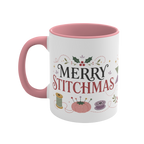 The left side of a white ceramic mug with a pink handle and interior, decorated with a bobbin, thread spool, pincushion and holly and the text ‘Merry Stitchmas’.