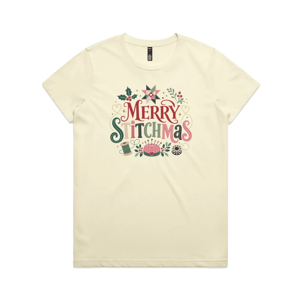A light butter yellow short-sleeve crewneck t-shirt featuring a colorful graphic print on the front that reads 'Merry Stitchmas' in decorative lettering with Christmas and quilting-themed illustrations.