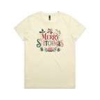 A light butter yellow short-sleeve crewneck t-shirt featuring a colorful graphic print on the front that reads 'Merry Stitchmas' in decorative lettering with Christmas and quilting-themed illustrations.