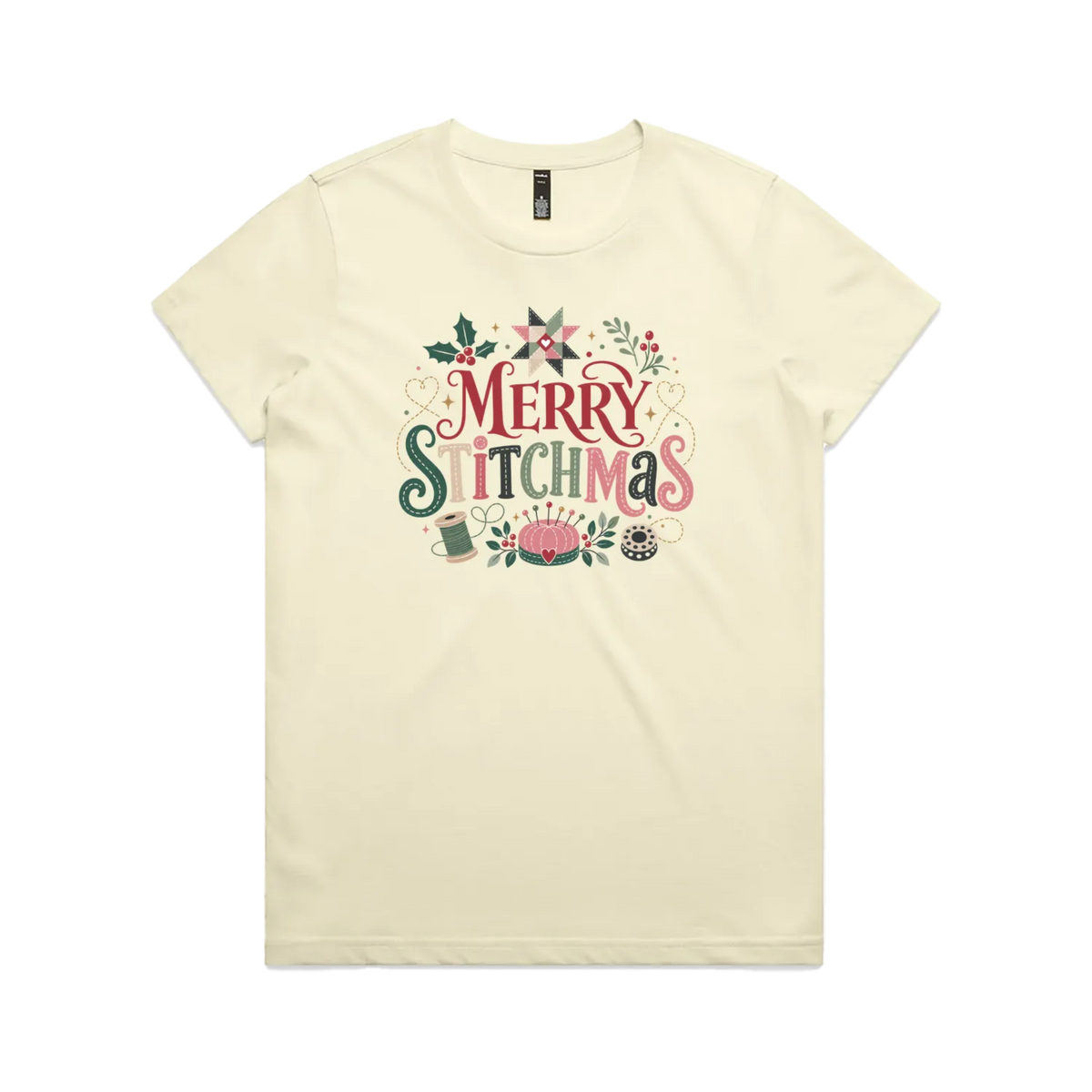 A light butter yellow short-sleeve crewneck t-shirt featuring a colorful graphic print on the front that reads 'Merry Stitchmas' in decorative lettering with Christmas and quilting-themed illustrations.