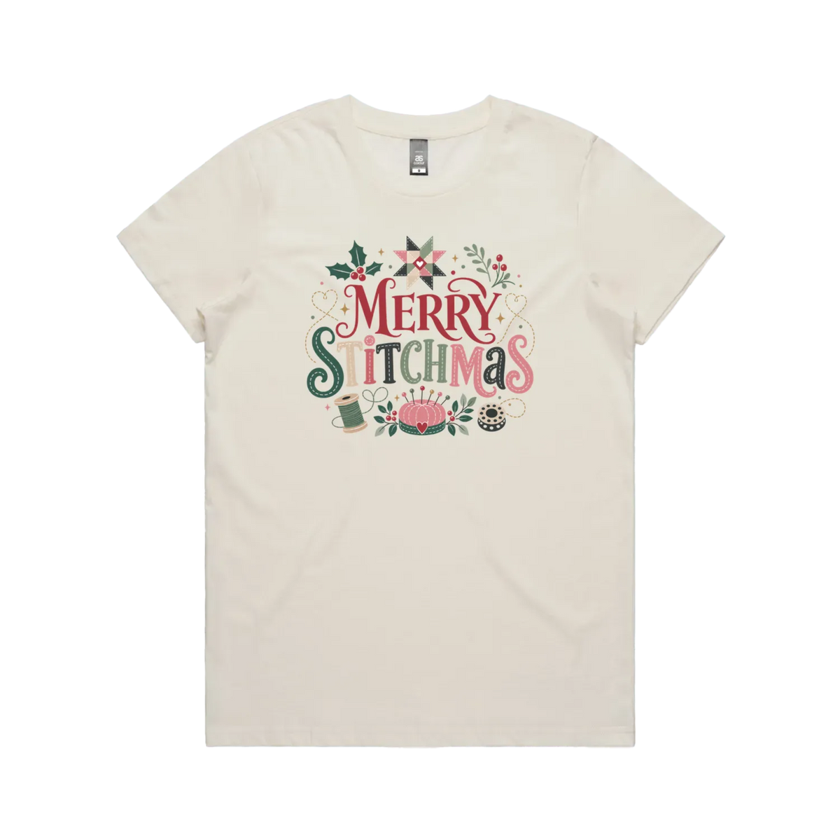 An off-white short-sleeve crewneck t-shirt featuring a colorful graphic print on the front that reads 'Merry Stitchmas' in decorative lettering with Christmas and quilting-themed illustrations.