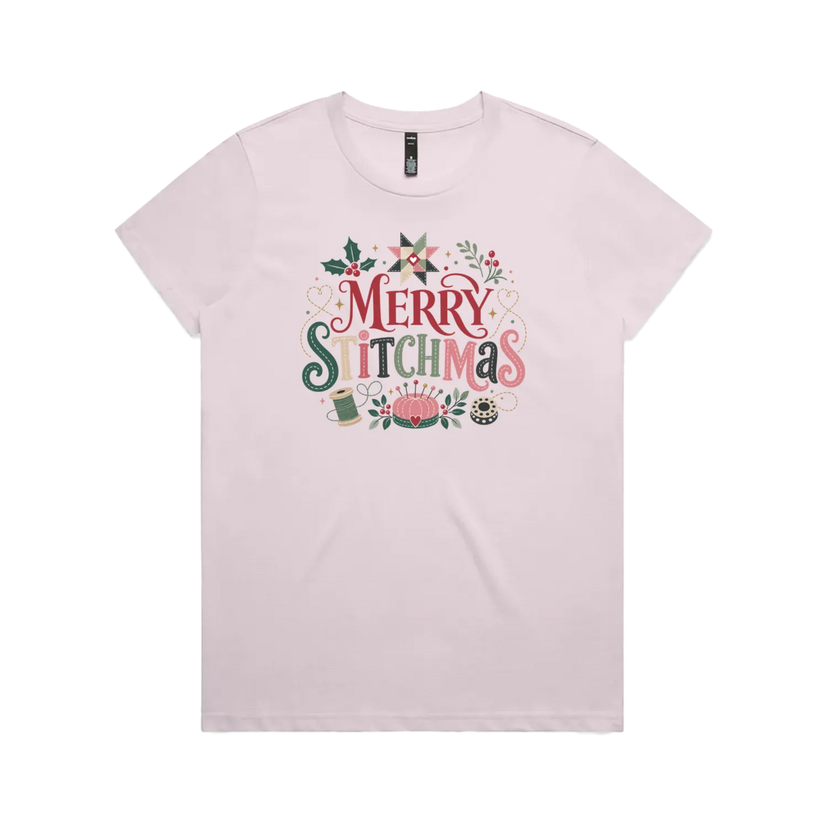 A light mauve short-sleeve crewneck t-shirt featuring a colorful graphic print on the front that reads 'Merry Stitchmas' in decorative lettering with Christmas and quilting-themed illustrations.