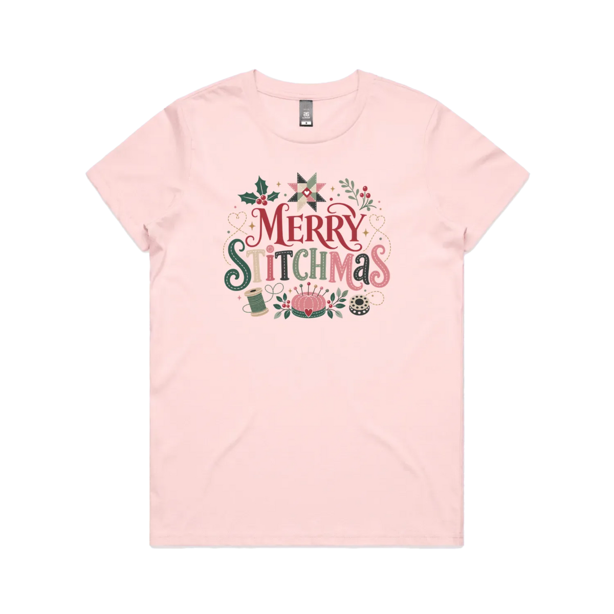 A light pink short-sleeve crewneck t-shirt featuring a colorful graphic print on the front that reads 'Merry Stitchmas' in decorative lettering with Christmas and quilting-themed illustrations.