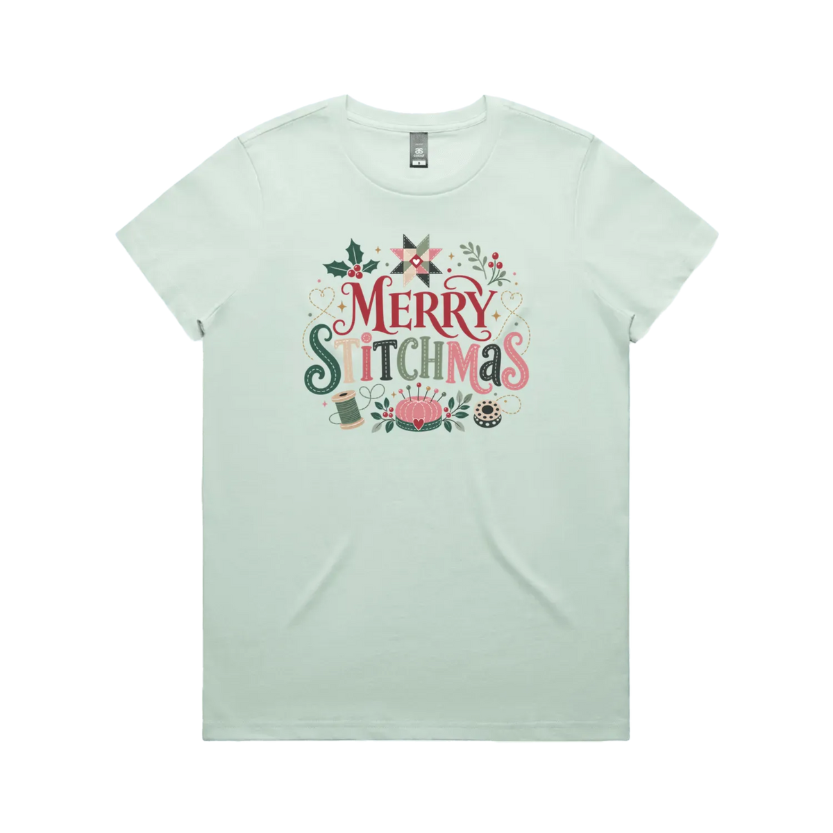 A light seafoam green short-sleeve crewneck t-shirt featuring a colorful graphic print on the front that reads 'Merry Stitchmas' in decorative lettering with Christmas and quilting-themed illustrations.