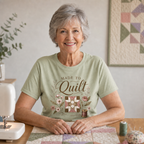 An older woman with gray hair sits at a table wearing a light pistachio green graphic t-shirt that reads 'Made to Quilt' with a quilt block, flowers and thread reel. She is surrounded by quilting items.