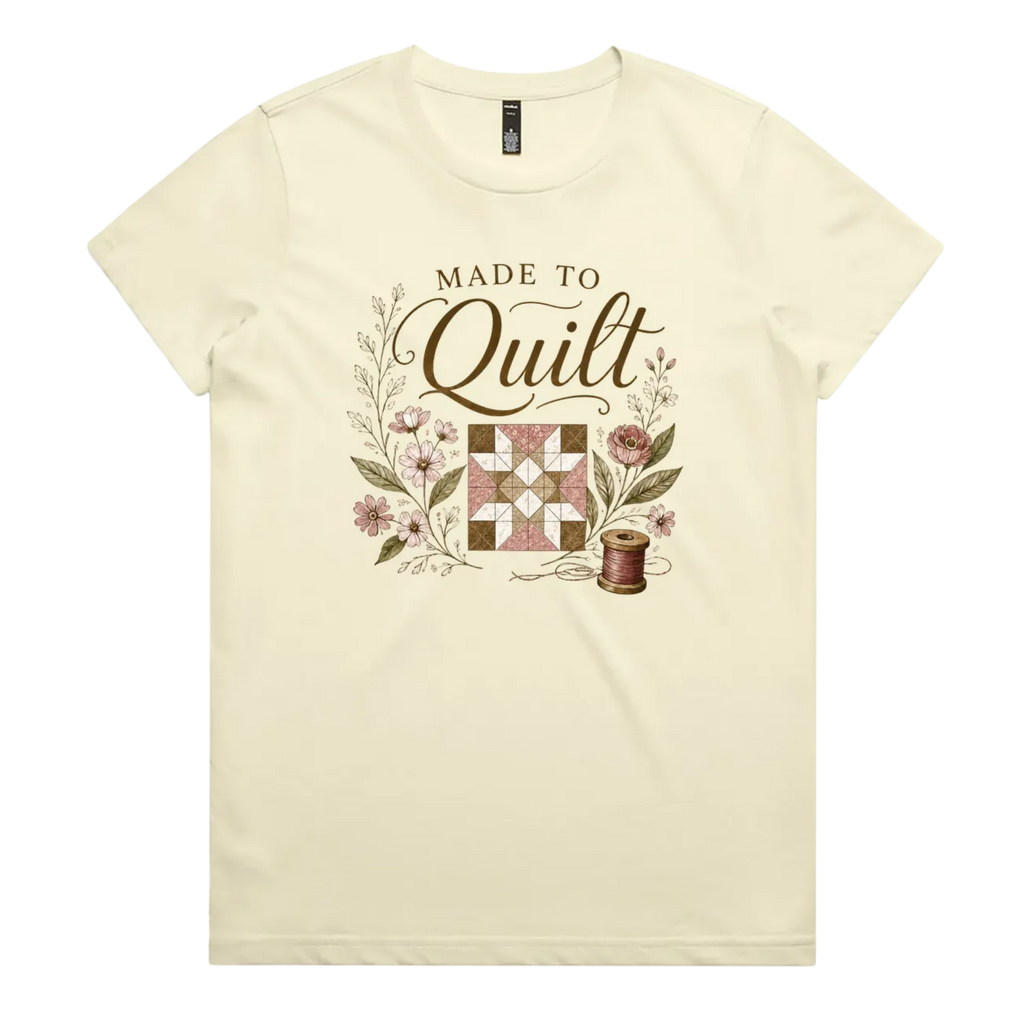 A very pale yellow short-sleeve crewneck t-shirt featuring a vintage-style quilt graphic with the text 'Made To Quilt' in brown script and floral illustrations.