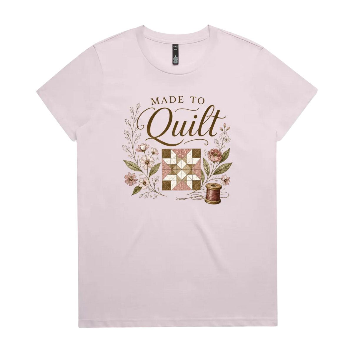 A light mauve short-sleeve crewneck t-shirt featuring a vintage-style quilt graphic with the text 'Made To Quilt' in brown script and floral illustrations.