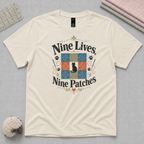 A light cream crewneck t-shirt with a colorful graphic print reading 'Nine Lives, Nine Patches' around a nine patch quilt block with a black cat silhouette in the middle. The shirt is displayed flat on a gray textured background with sewing props.