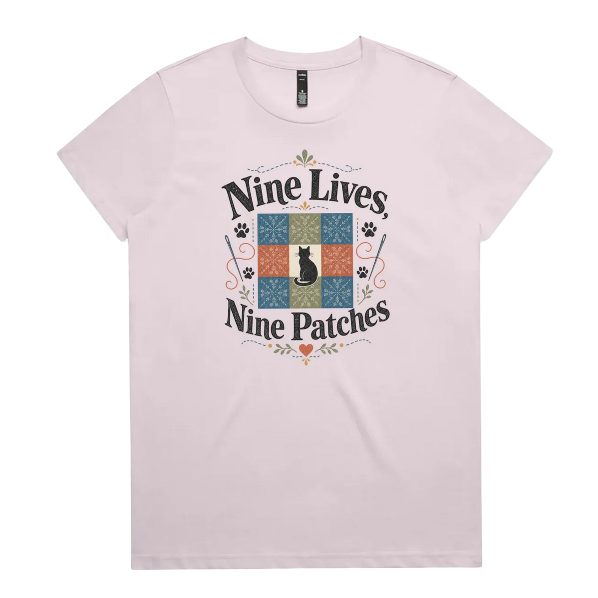 A light mauve short-sleeve t-shirt with a colorful graphic on the front that reads 'Nine Lives, Nine Patches' around a nine patch quilt block with a black cat silhouette in the middle.