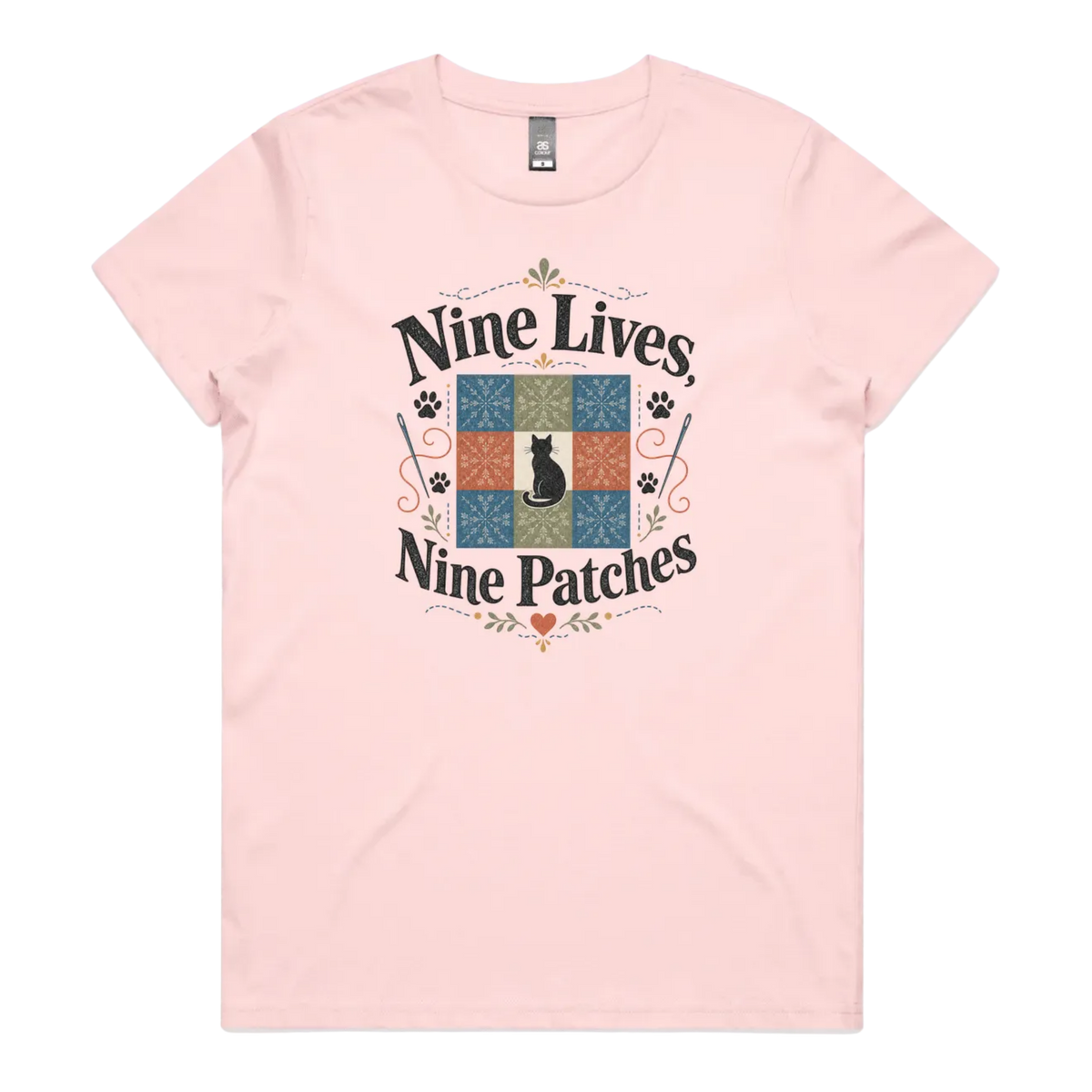 A light pink short-sleeve t-shirt with a colorful graphic on the front that reads 'Nine Lives, Nine Patches' around a nine patch quilt block with a black cat silhouette in the middle.