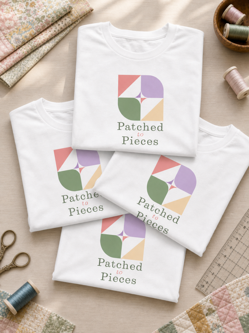 Three folded white T-shirts arranged on a light wood surface, each printed with a colorful geometric patchwork logo and the words 'Patched to Pieces.' Quilted fabric, spools of thread, scissors, and a ruler are styled around them.