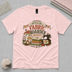 A light pink t-shirt with the words 'Official Fabric Hoarder Society' above a stack of fabric, a sewing machine and mushrooms. It is laid flat on a light grey cutting mat with sewing supplies partially visible in the corners.