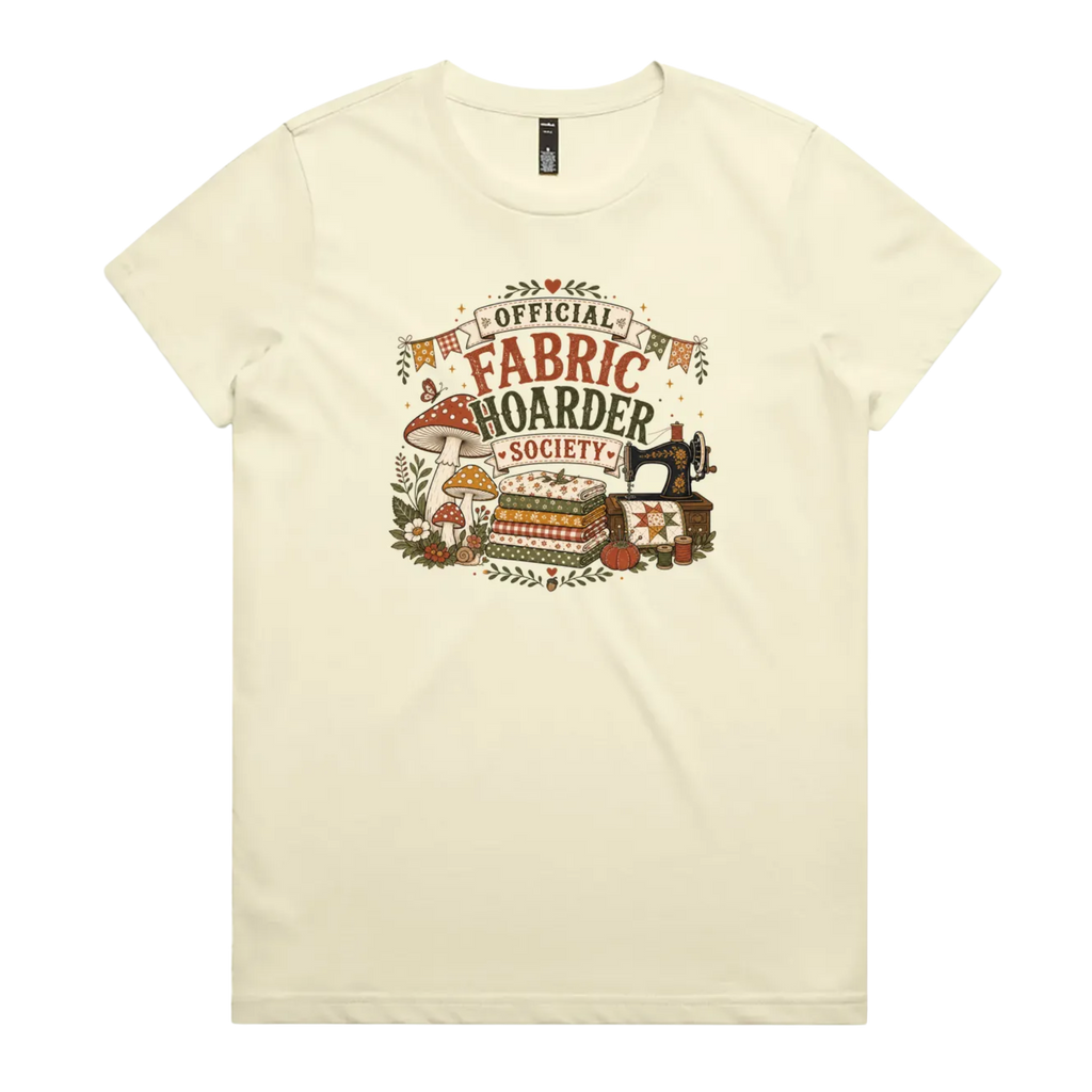 A pale butter yellow crewneck short-sleeved t-shirt with the words 'Official Fabric Hoarder Society' above an illustration of a stack of fabric, a sewing machine and a group of muchrooms, flowers and a snail.