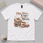 A white short-sleeve graphic T-shirt laid flat on a light grey cutting mat. The shirt features a vintage-style sewing and quilting illustration with the text 'One More Cup, One More Block' in brown and black lettering.