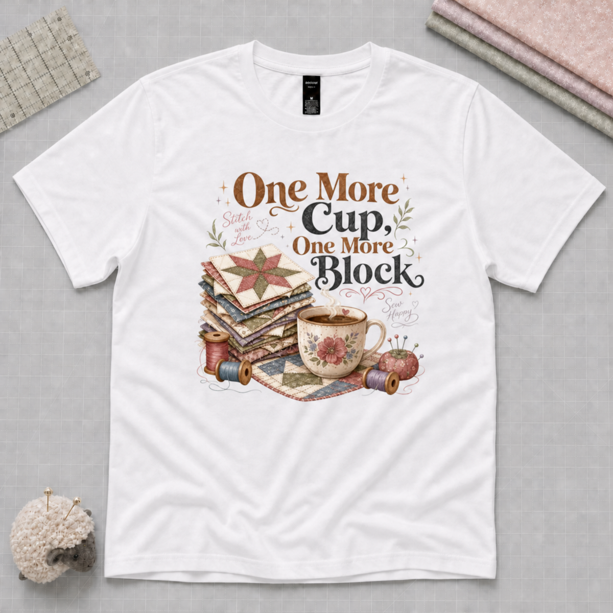 A white short-sleeve graphic T-shirt laid flat on a light grey cutting mat. The shirt features a vintage-style sewing and quilting illustration with the text 'One More Cup, One More Block' in brown and black lettering.