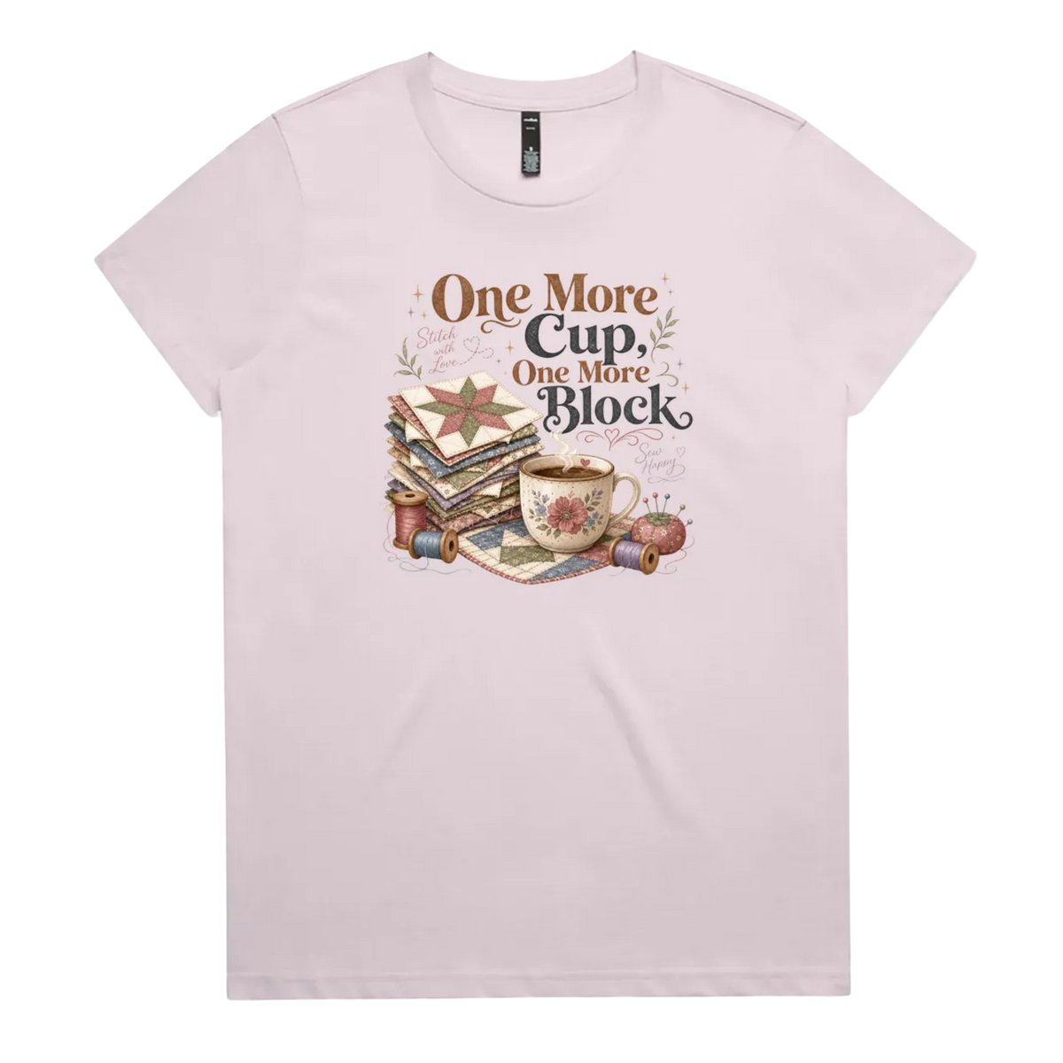 A pale mauve short-sleeve crewneck t-shirt featuring a vintage-style graphic of stacked quilt blocks, a coffee cup, and thread spools with the text 'One More Cup, One More Block' in brown and black lettering.