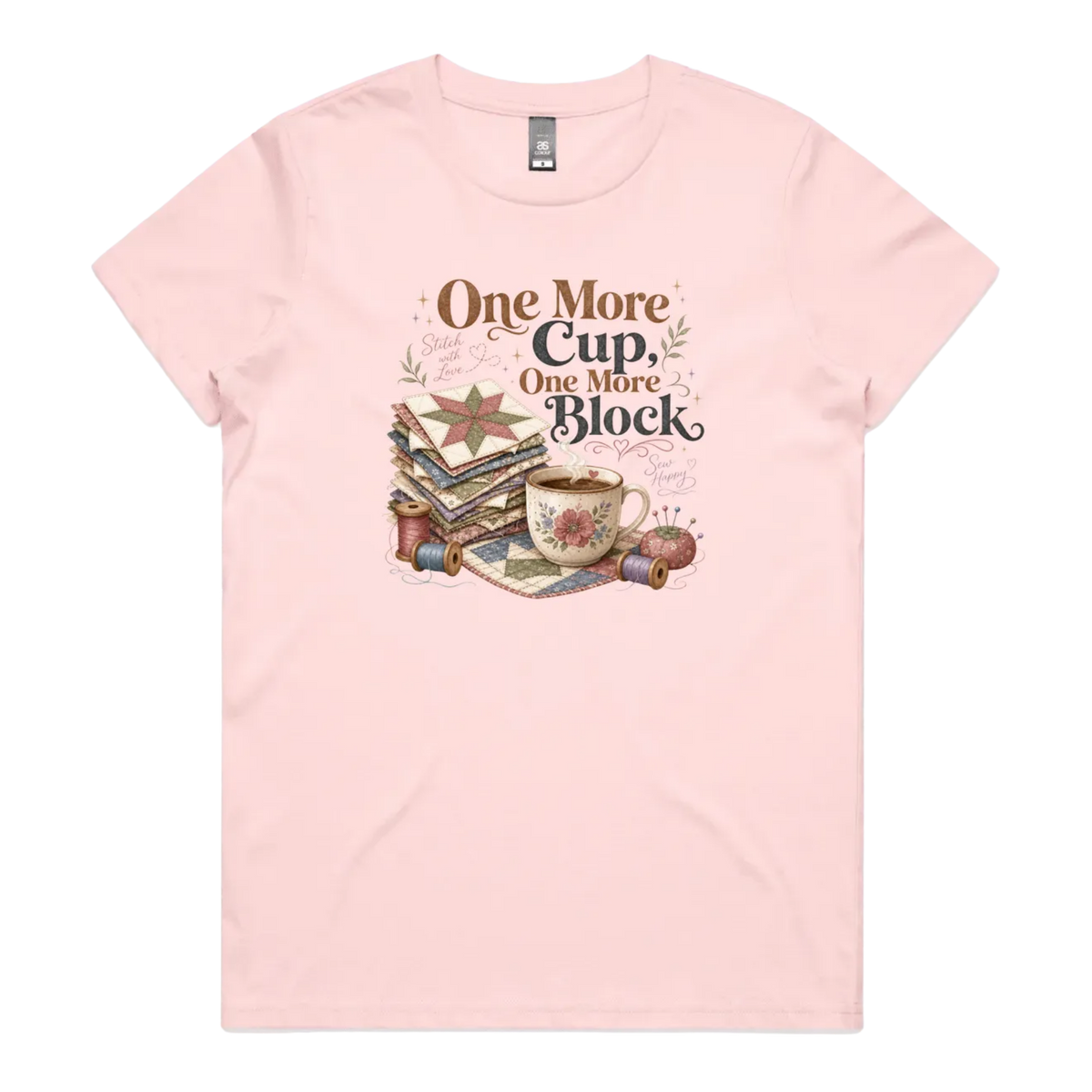 A light pink short-sleeve crewneck t-shirt featuring a vintage-style graphic of stacked quilt blocks, a coffee cup, and thread spools with the text 'One More Cup, One More Block' in brown and black lettering.