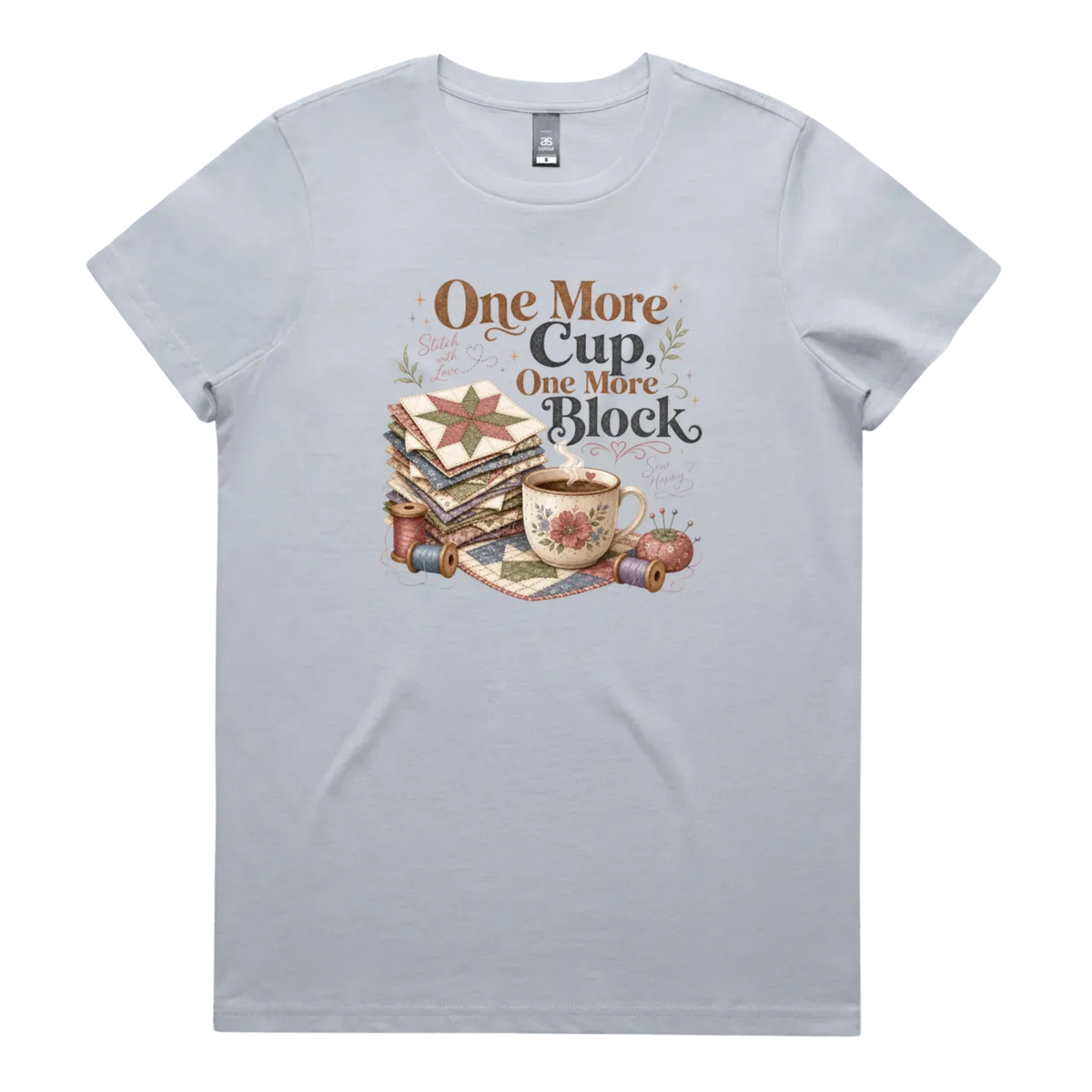 A light powder blue short-sleeve crewneck t-shirt featuring a vintage-style graphic of stacked quilt blocks, a coffee cup, and thread spools with the text 'One More Cup, One More Block' in brown and black lettering.