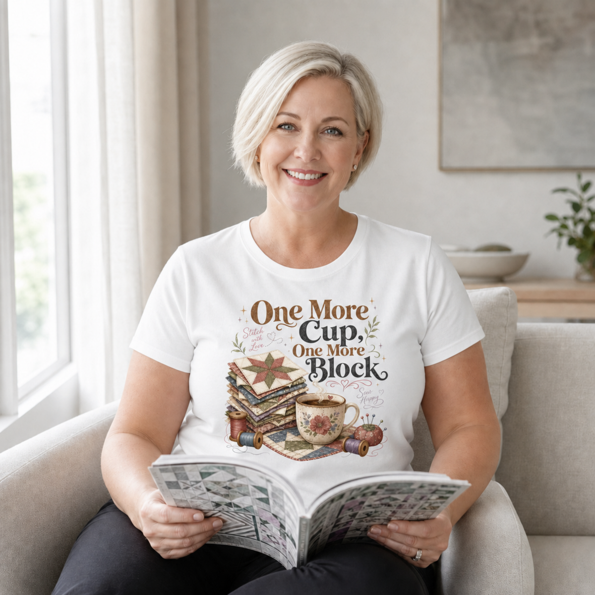 A woman with short blond hair sitting on a beige lounge holding an open book. She is wearing a white t-shirt featuring the words 'One More Cup, One More Block' above an illustration of a coffee cup, quilt blocks, thread spools and a pincushion.