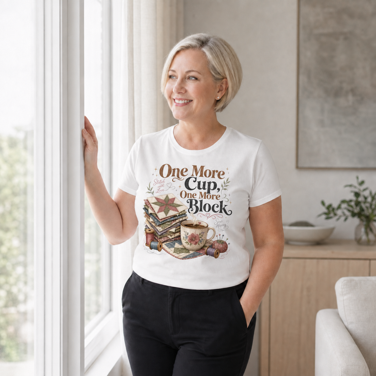 A smiling woman with short blonde hair stands by a window wearing a white graphic T-shirt that reads 'One More Cup, One More Block' above an illustration of a coffee cup, quilt blocks, thread spools and a pincushion.
