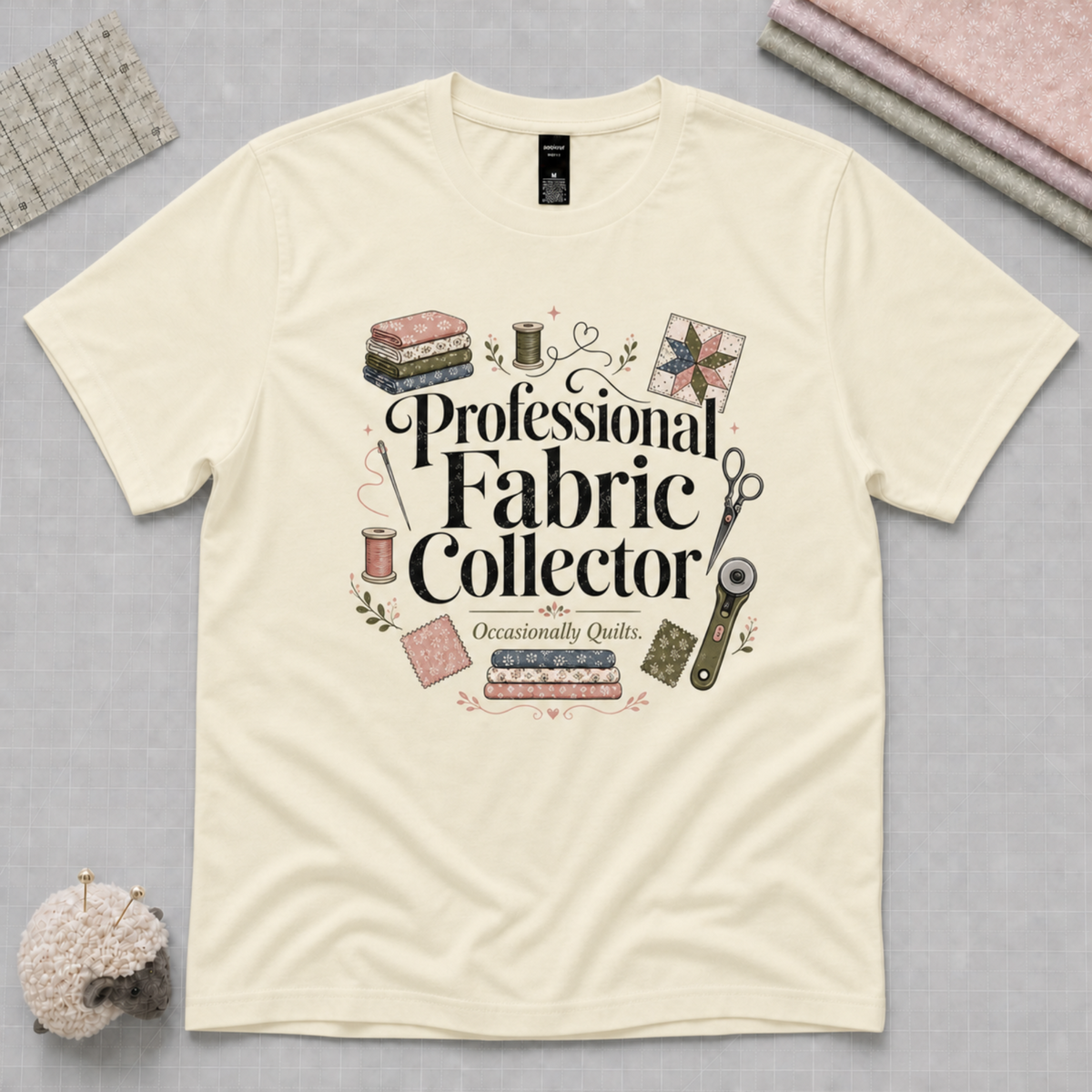 A pale butter yellow short-sleeve t-shirt with a 'Professional Fabric Collector' graphic print, styled in a flat-lay arrangement with sewing-themed props on a gray background.