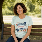A smiling woman with short brown hair sits on a wooden park bench wearing a pale mint green graphic t-shirt. The t-shirt features the text 'Perfectly Imperfect Stitches' in blue and pink script, surrounded by sashiko-themed illustrations like thread spools, scissors, and fabric squares.