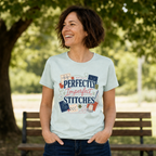A woman smiling outdoors in a park, wearing a light mint green graphic t-shirt with the text 'Perfectly Imperfect Stitches' in a colourful patchwork design, paired with dark blue jeans.