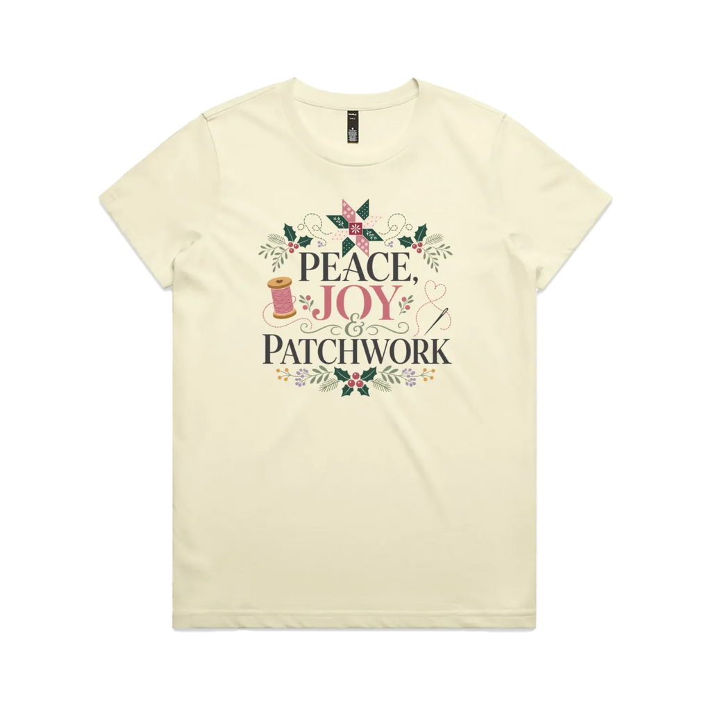 A pale butter yellow short-sleeve crewneck t-shirt with a central graphic print that reads 'Peace, Joy & Patchwork' in dark lettering, decorated with a pink bow, quilt block, spool of thread, needle, and floral elements.