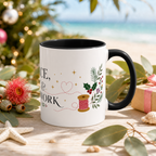 The right side of a white ceramic mug with a black handle and interior, decorated with a thread spool, stitch lines, holly, berries and flowers. The text ‘Peace, Joy & Patchwork' is partially visible. The mug is styled on a wooden surface with eucalyptus leaves, a starfish, a pink flower, and a blurred beach and Christmas tree background.