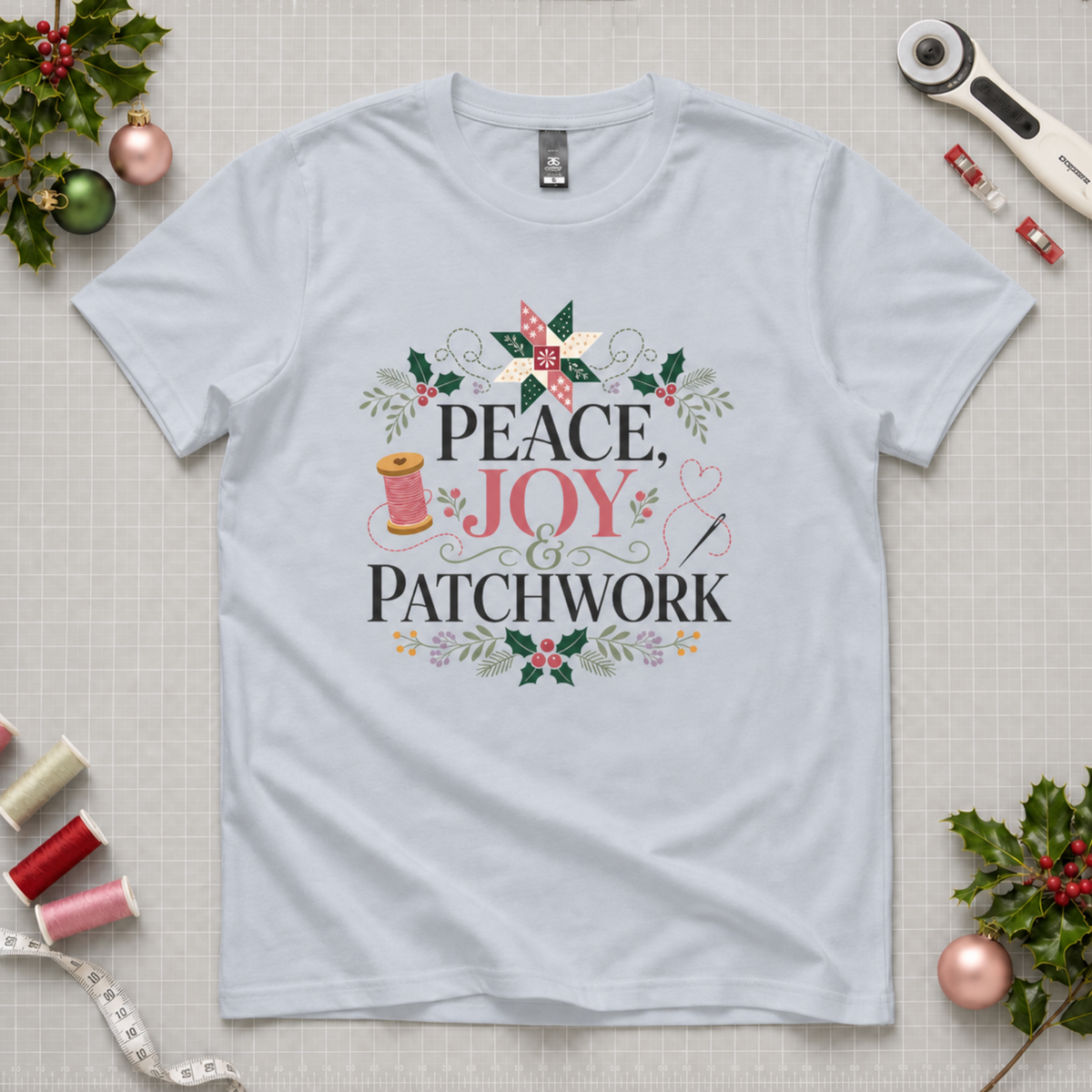 A powder blue crewneck T-short laid flat on a grey cutting mat with sewing equipment and holly in the corners. The graphic on the tee reads 'Peace, Joy & Patchwork' surrounded by a patchwork star, thread, needle holly and flowers.