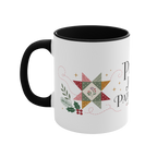 The left side of a white ceramic mug with a black handle and interior, decorated with a quilt block, stitch lines and holly. The text ‘Peace, Joy & Patchwork' is partially visible.