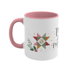 The left side of a white ceramic mug with a pink handle and interior, decorated with a quilt block, stitch lines and holly. The text ‘Peace, Joy & Patchwork' is partially visible.