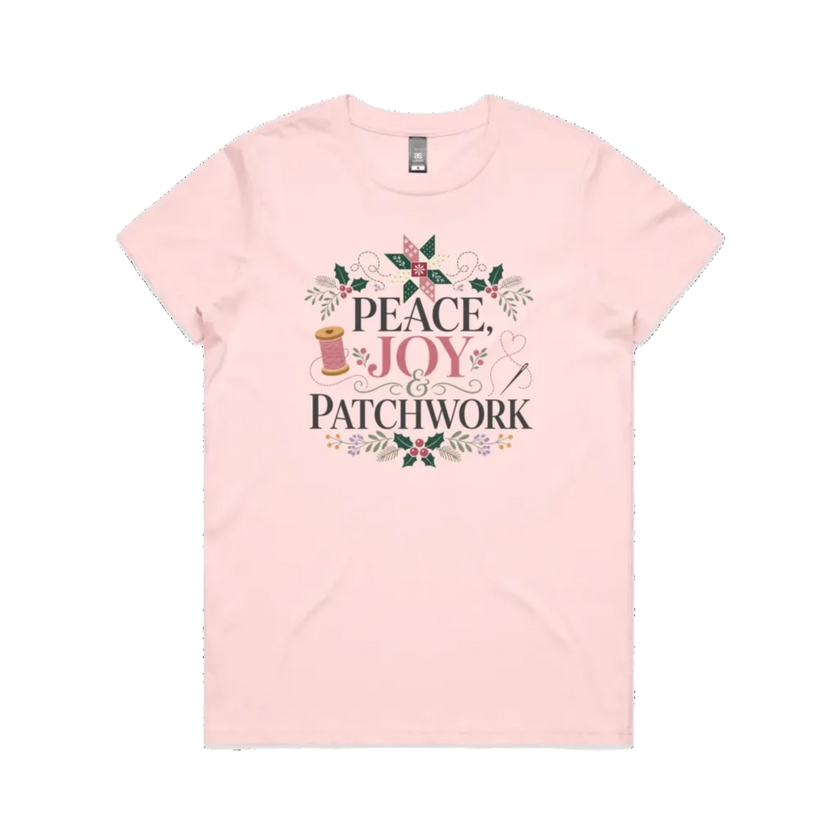 A light pink short-sleeve crewneck t-shirt with a central graphic print that reads 'Peace, Joy & Patchwork' in dark lettering, decorated with a pink bow, quilt block, spool of thread, needle, and floral elements.