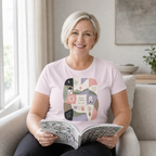 A smiling woman with short blonde hair sits in a light gray armchair, holding an open book. She is wearing a light mauve crewneck t-shirt with a colorful, whimsical graphic on the front that reads 'Pieces of Home'.