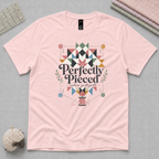 A pale pink short-sleeve graphic t-shirt with the words 'Perfectly Pieced Modern Quilt Guild' cutting through a patchwork star. It lays flat on a grey background with quilting props in the corners.