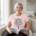 A blond smiling woman sitting on a beige lounge holding an open book. She is wearing a pink short-sleeve t-shirt with the graphic 'Perfectly Pieces Modern Quilt Guild' written across a large patchwork star.