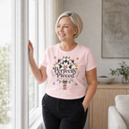 A blond smiling woman standing at a window wearing a pink short-sleeve t-shirt with the graphic 'Perfectly Pieces Modern Quilt Guild' written across a large patchwork star.