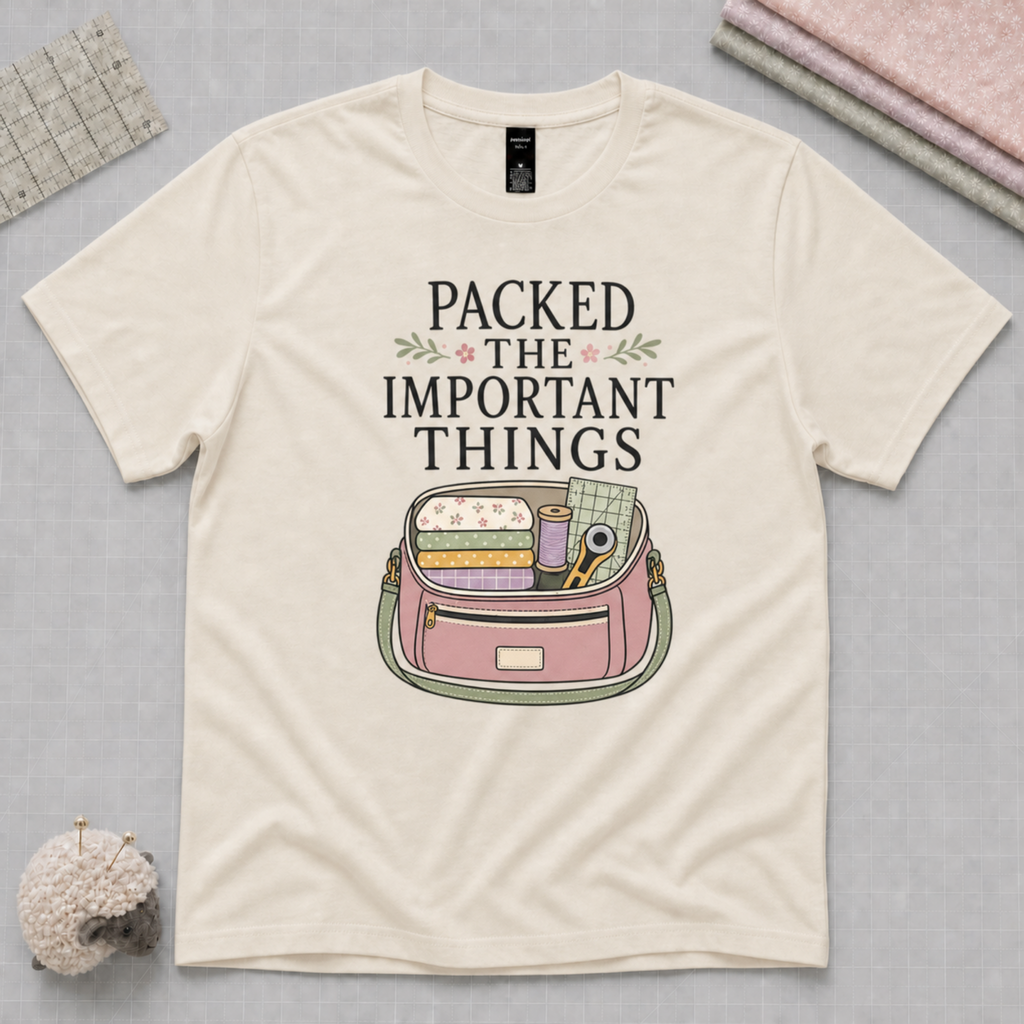 An off-white short-sleeve t-shirt featuring the words 'Packed The Important Things' above an image of a small bag containing sewing supplies. The shirt lays flat on a pale grey cutting mat with a quilting ruler, folded fabric and pincushion in the corners.