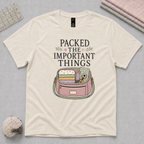 An off-white short-sleeve t-shirt featuring the words 'Packed The Important Things' above an image of a small bag containing sewing supplies. The shirt lays flat on a pale grey cutting mat with a quilting ruler, folded fabric and pincushion in the corners.