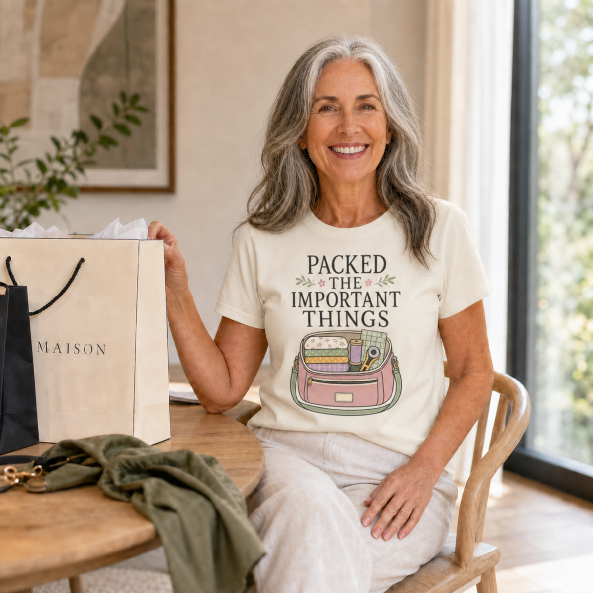 A smiling woman with gray hair sits indoors at a wooden table, wearing a cream graphic T-shirt that reads 'PACKED THE IMPORTANT THINGS' above a pink suitcase illustration. She holds a large shopping bag beside her, with a green jacket on the table and a bright room with large windows in the background.