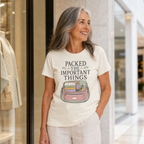 A smiling older woman with gray hair stands in a shopping mall next to a storefront, wearing a cream-colored graphic t-shirt that reads 'PACKED THE IMPORTANT THINGS' above a pink and green suitcase illustration.